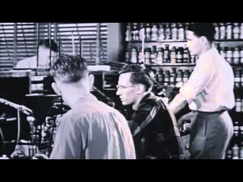 This image is black and white shows three guys sitting at a table and what looks like some type of manufacturing plant. On the table it looks like some type of chemical experimenting or formulating going on. This image connects to a video so just click and it will play.